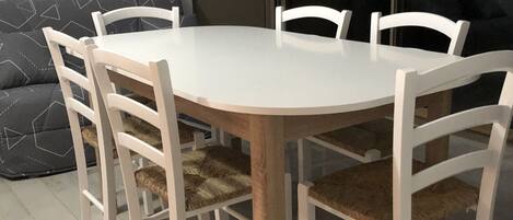 Une table de salle à manger ronde, entourée de chaises blanches, un canapé gris et une grande fenêtre.