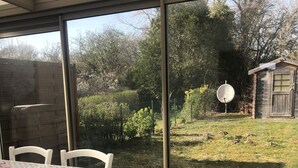Een overdekt terras met een eettafel, grote schuifdeuren en uitzicht op een tuin en een klein houten gebouw.