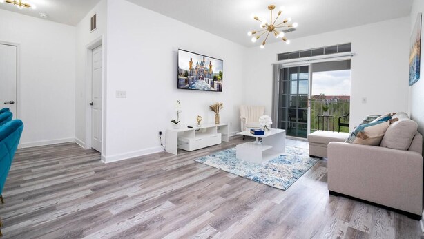 A modern living room with a white sofa, a coffee table, and a TV mounted on the wall. There is a large window providing a view of the outdoors. - 2BR Mickey Magic Condo | Family Stay Near Disney + Pool (Kissimee)