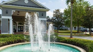 A building with a fountain in front, surrounded by greenery and a clear sky. - 2BR Mickey Magic Condo | Family Stay Near Disney + Pool (Kissimee)