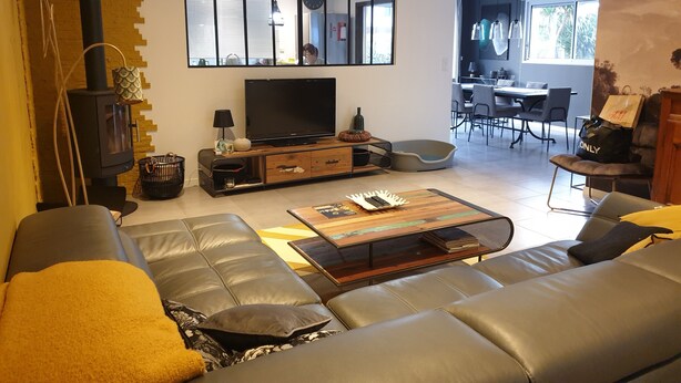 A modern living room with a large sofa, a TV, a fireplace, and a dining area visible through a glass partition. - 3-star Villa Nelson sleeps 6 3 bedrooms private pool (Moliets-et-Maa)