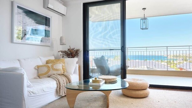 A modern living room with a glass coffee table, white sofa, and a view of the sea through a sliding glass door.