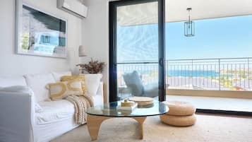 A modern living room with a glass coffee table, white sofa, and a view of the sea through a sliding glass door.