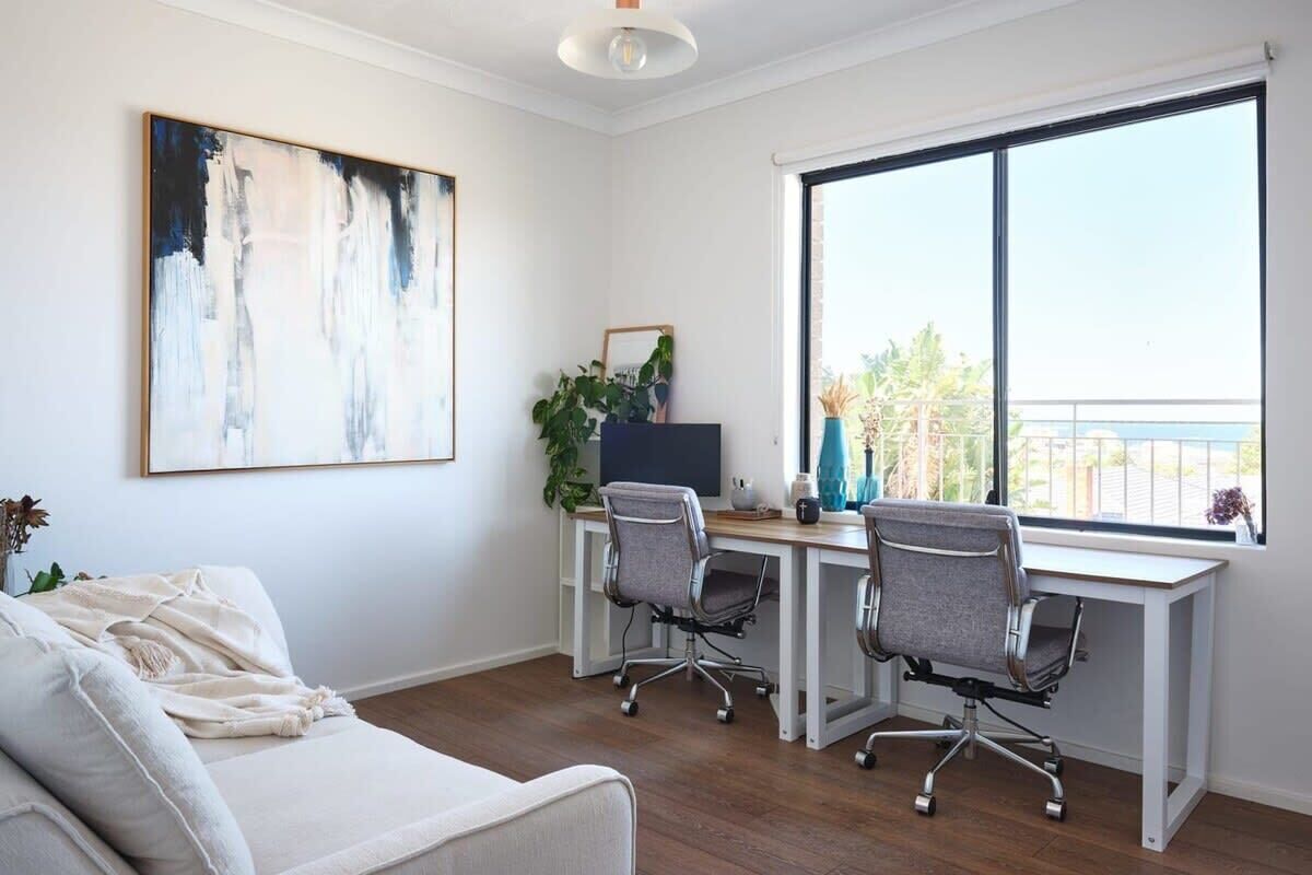 A modern office space with a white couch, two office chairs, a desk, and a large window with a view of greenery.