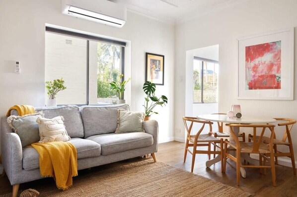 A modern living room with a grey sofa, wooden dining table, and chairs. There are two windows with plants on either side, and a framed picture on the wall.