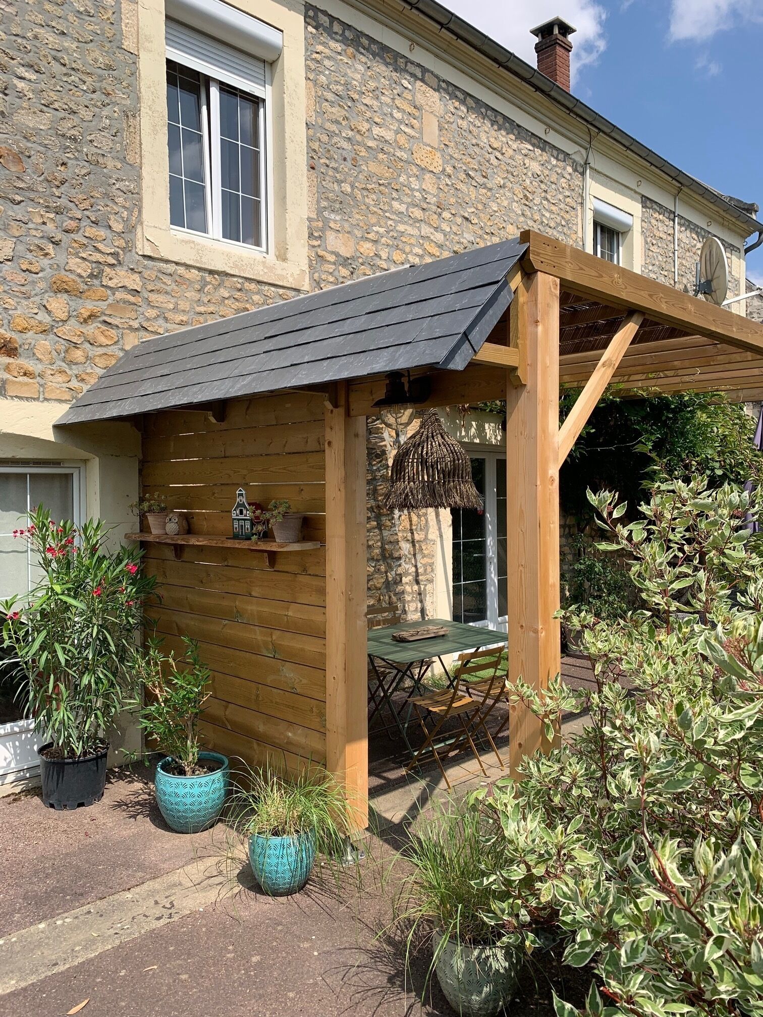 A wooden pergola with a sloped roof, a stone wall, and a table with chairs underneath.
