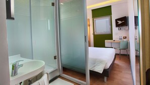 A modern hotel room with a glass door leading to a shower area, a bed with white bedding, a small desk with a chair, and a mounted TV. - Hotel Caspia Pro (Greater Noida)