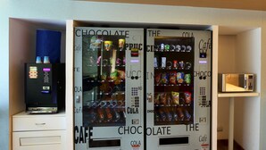 A room with a minibar, a coffee machine, and two vending machines labeled "Chocolate" and "The Cappuccino." - Hotel Caspia Pro (Greater Noida)