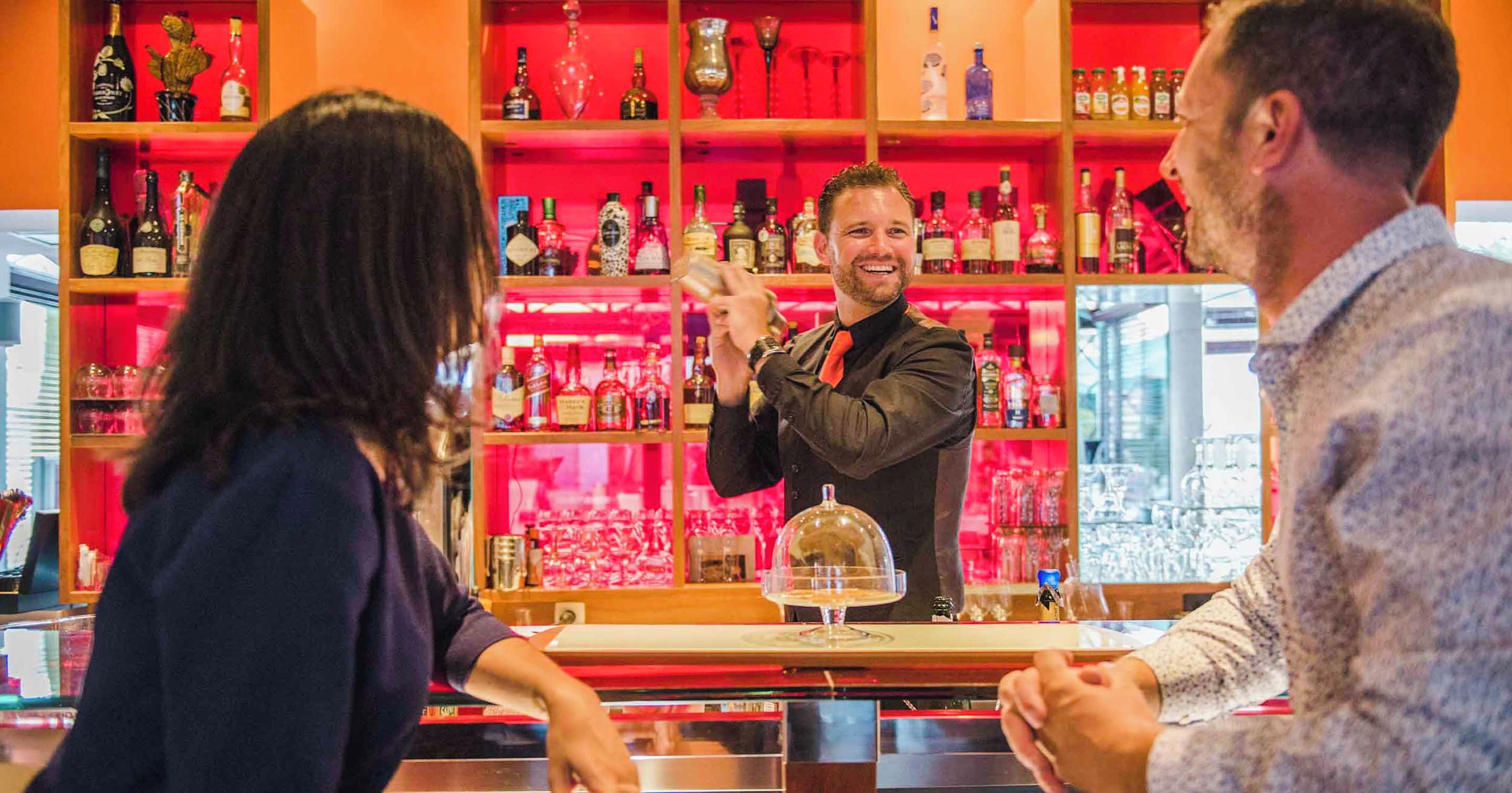 A bar area with shelves of bottles and glasses, a bartender, and customers.