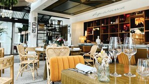 L’intérieur d’un restaurant présente des chaises en osier, une table en bois avec des verres à vin et un coin bar avec des étagères remplies de bouteilles.