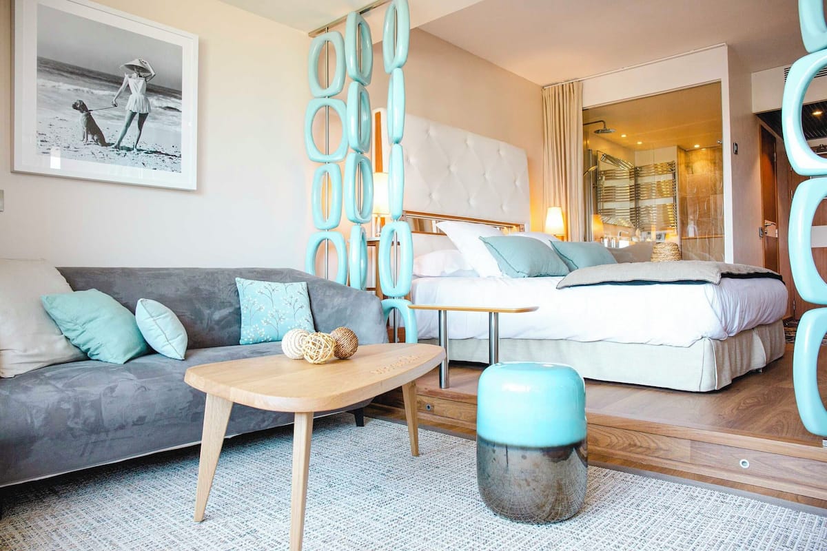 Ein modernes Hotelzimmer mit grauem Sofa, einem Holztisch, einem Bett mit weißem Kopfteil und einem durchgegangene Tür erkennbaren Badezimmer.