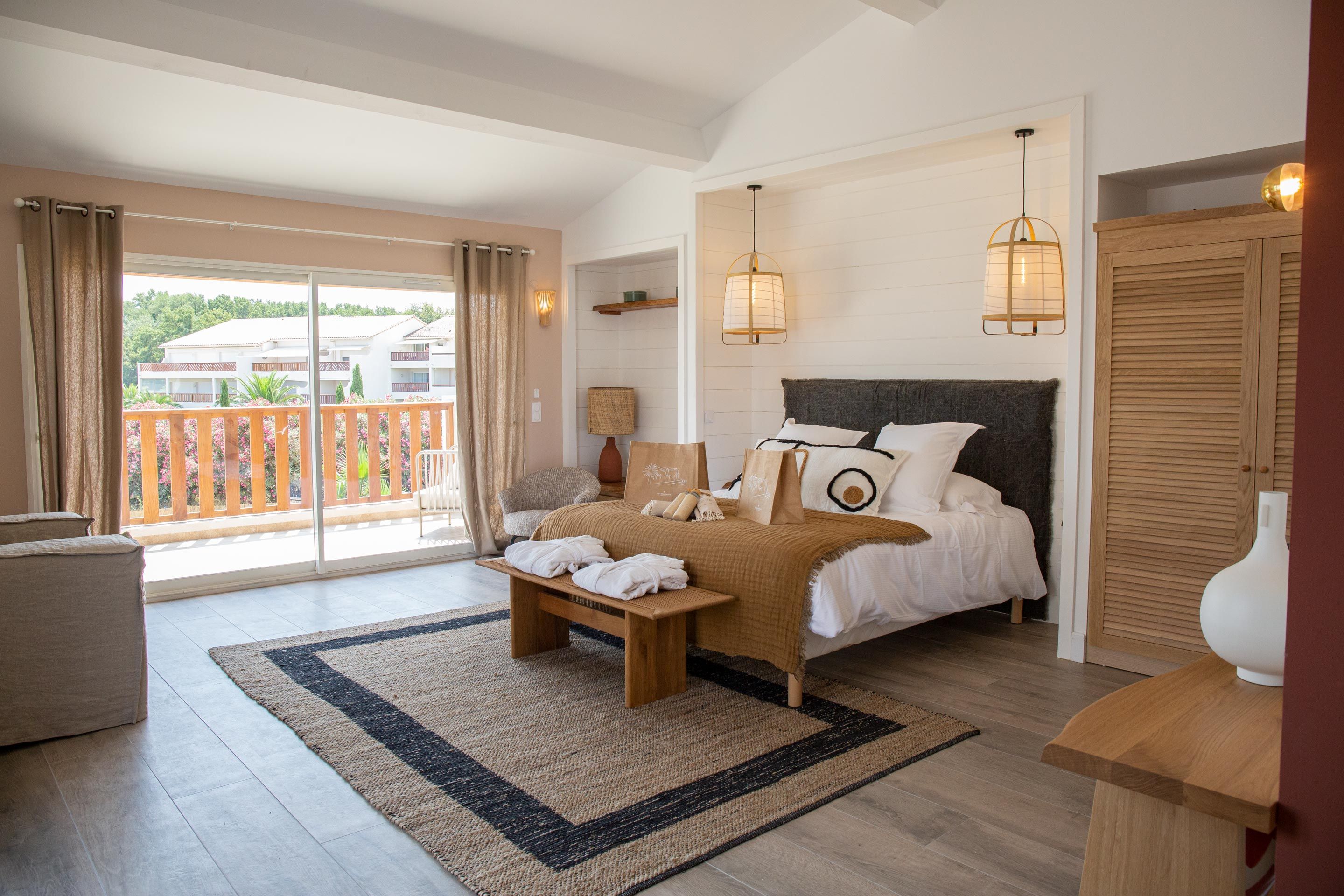 A bedroom with a bed, a bench, a bedside table, a lamp, and a view of the outside.