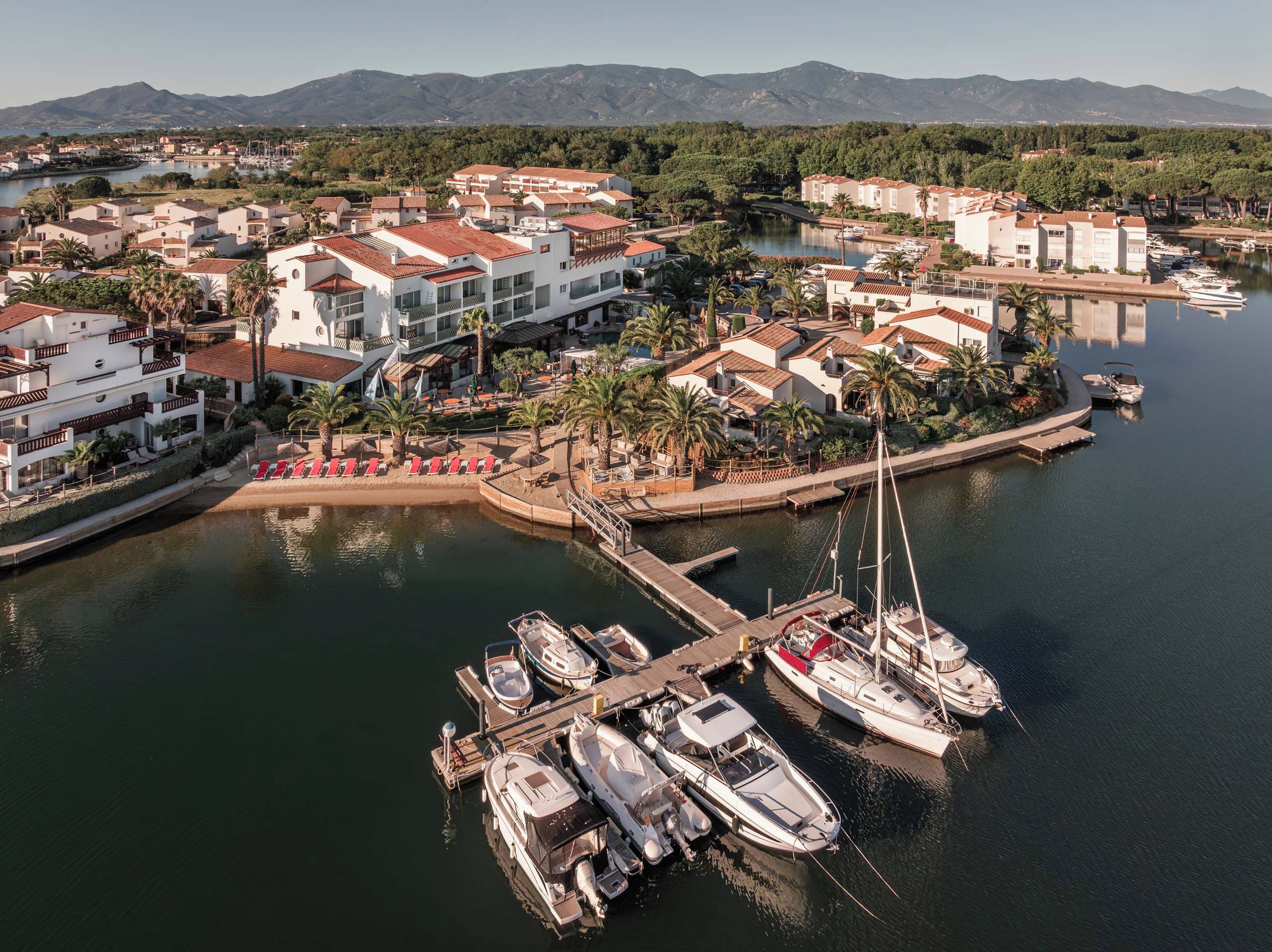 Un puerto deportivo con barcos amarrados, rodeado de edificios con tejados rojos y palmeras.