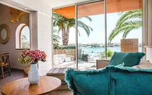 Een moderne kamer met een grote schuifglazen deur die uitkomt op een balkon met uitzicht op zee en palmbomen. Er staat een rond houten tafeltje met een bloevas en een bank met een blauwgroen sierkussen.