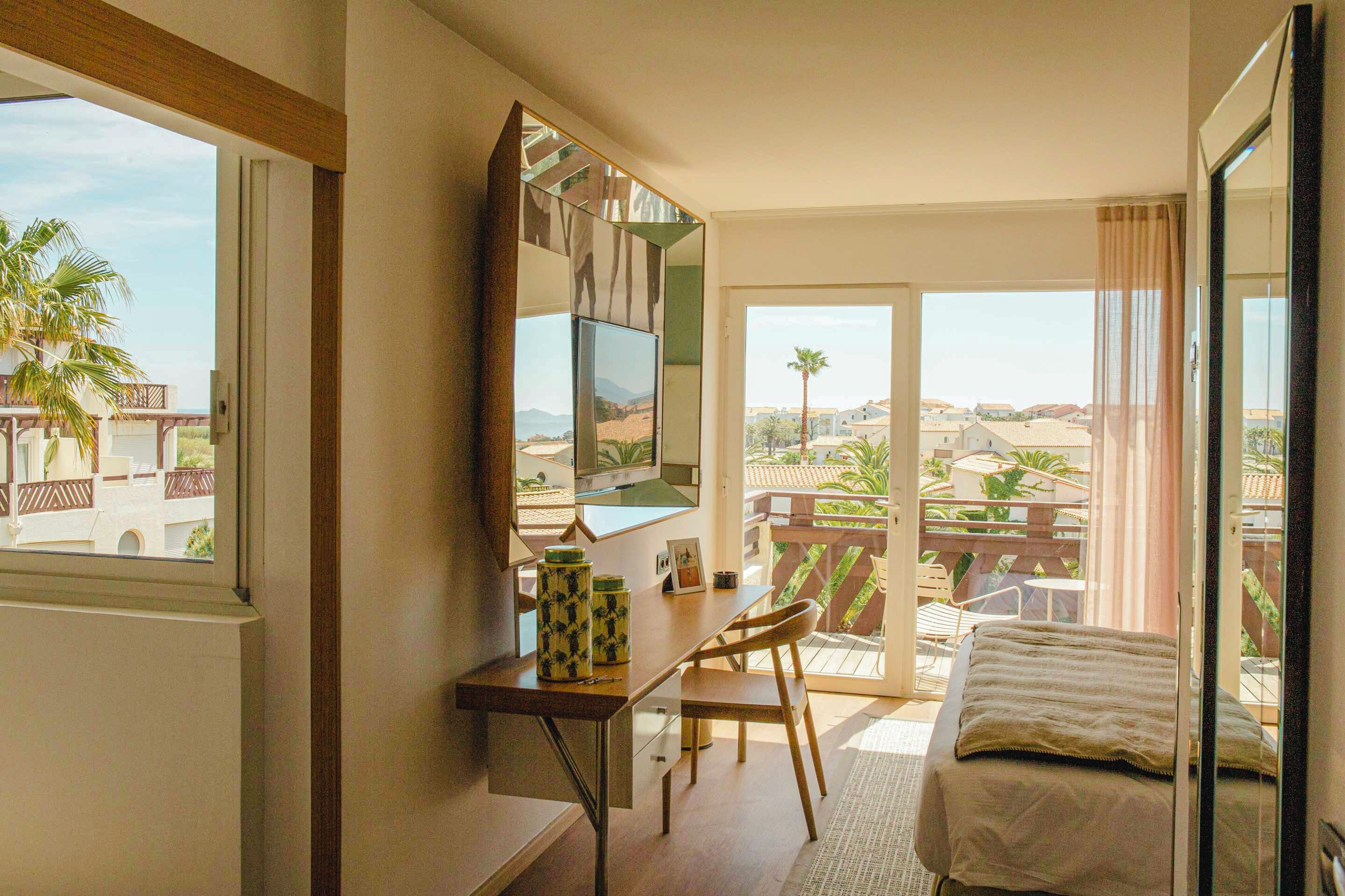 A hotel room with a desk, chair, bed, and a balcony with a view of palm trees and buildings.