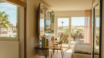 A hotel room with a desk, chair, bed, and a balcony with a view of palm trees and buildings.