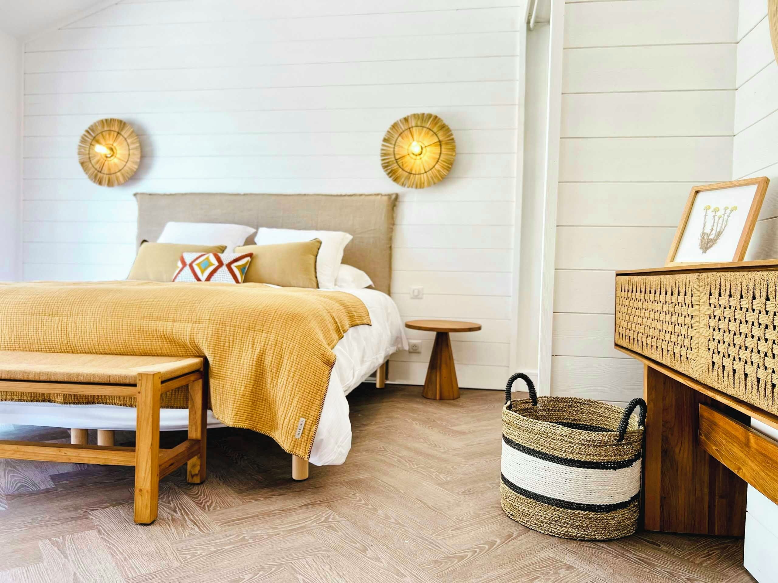 A bedroom with a bed, bedside tables, a bench, a woven basket, and wall-mounted lamps.
