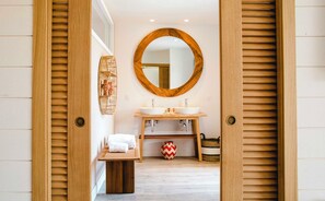 Ein Badezimmer mit Holztür, rundem Spiegel, doppelter Waschbecken und kleinem Tisch mit Handtüchern.