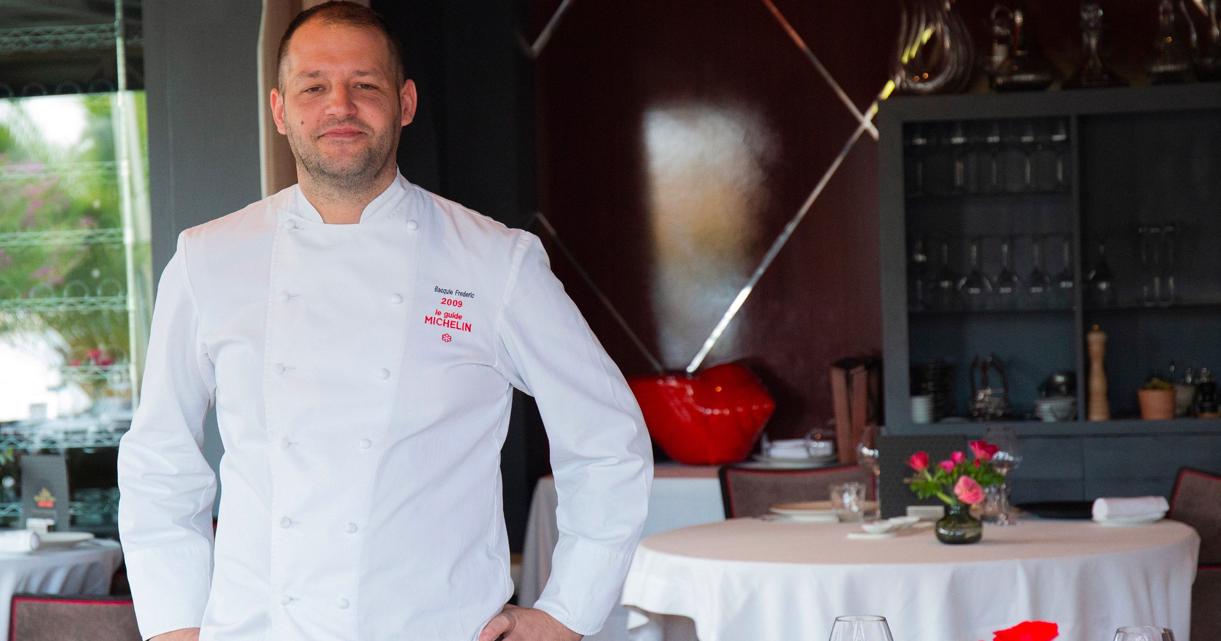 A chef in a white uniform standing in a restaurant with a round table, chairs, and a red decorative object in the background.