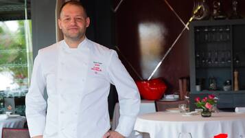 A chef in a white uniform standing in a restaurant with a round table, chairs, and a red decorative object in the background.
