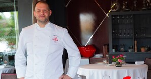 Un chef, vêtu d’un uniforme blanc, se tient dans un restaurant. On y voit une table ronde, des chaises et, au fond, un objet décoratif rouge.