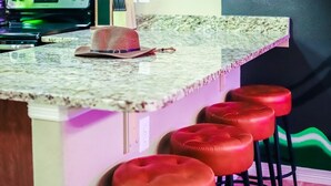 A kitchen counter with a woven hat and a pair of sunglasses, a granite countertop, and three red bar stools. - The Neon Cowboy: Hot Tub, Fire Pit, Movie Theater (Denver)