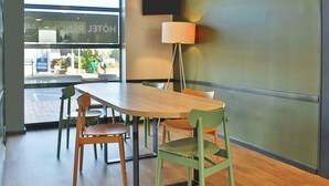 A hotel room with a wooden table, two chairs, a floor lamp, and a large window with a view of the street. - B&B Hotel Les Sables d Olonne Centre Gare (Les Sables-d'Olonne)