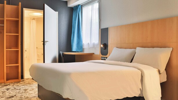 Une chambre d’hôtel avec un grand lit, une tête de lit en bois, un bureau avec une chaise, une armoire et une salle de bain visible à travers une porte ouverte.