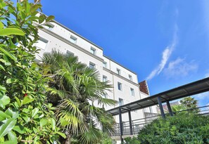 Un immeuble à plusieurs étages, doté d’une façade blanche et entouré d’une végétation luxuriante, notamment de palmiers et d’autres plantes.