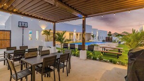 An outdoor patio with a pool, wooden tables and chairs, and a covered area with string lights. - Stunning 6-Bedroom Villa in Aruba (Noord)