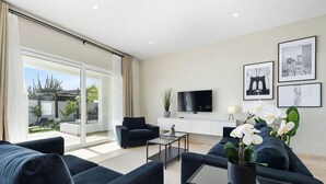 A modern living room with a sofa, coffee table, and TV. There is a balcony with plants and a cityscape view. - Stunning 6-Bedroom Villa in Aruba (Noord)