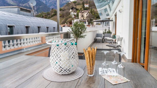 A wooden table with a white woven basket, a glass vase with breadsticks, and wine glasses on a balcony overlooking a mountainous landscape.