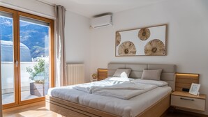 A modern bedroom with a large bed, a bedside table, a wall-mounted air conditioner, and a view of mountains through a sliding glass door.