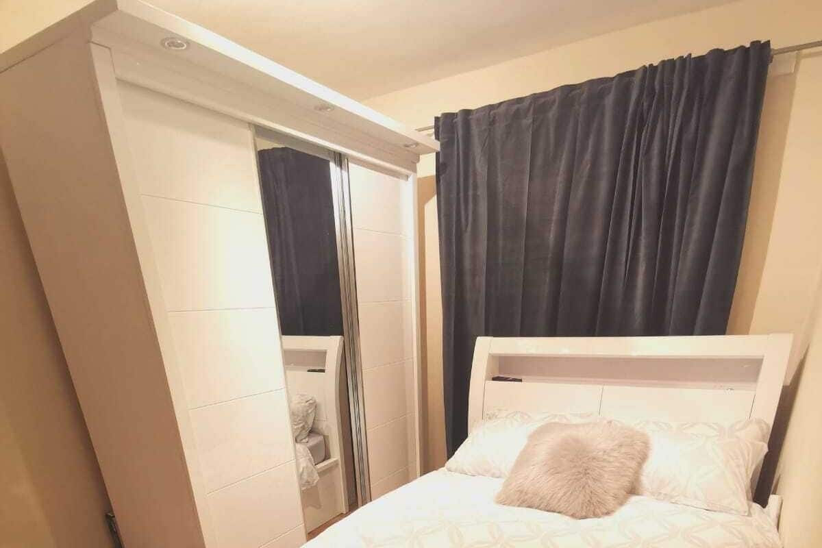 A bedroom with a bed, a small table, a chair, a trash bin, and a bathroom with a toilet and sink.