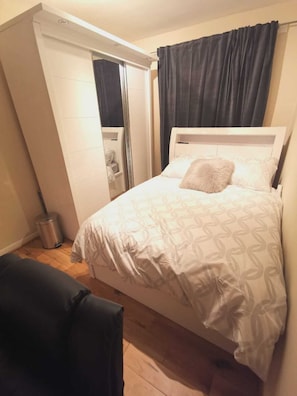 A bedroom with a bed, a small table, a chair, a trash bin, and a bathroom with a toilet and sink. - Umbrella Properties Woolwich London (London)