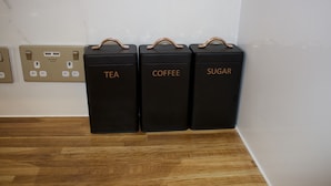 Three black containers labeled tea, coffee, and sugar, placed on a wooden floor next to a wall with electrical outlets. - Umbrella Properties Woolwich London  (London)