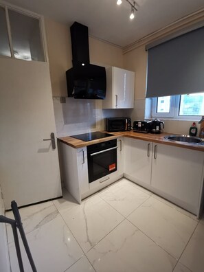 A kitchen with white cabinets, a black range hood, a microwave, a coffee maker, and a window with blinds. - Umbrella Properties Woolwich London (London)