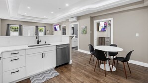 A modern kitchen with white cabinets, a stainless steel dishwasher, and a dining area with a round table and black chairs.