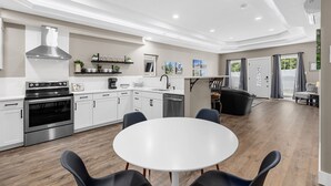 A modern kitchen with white cabinetry, a stainless steel range hood, and a built-in oven. There is a dining area with a round table and four chairs.