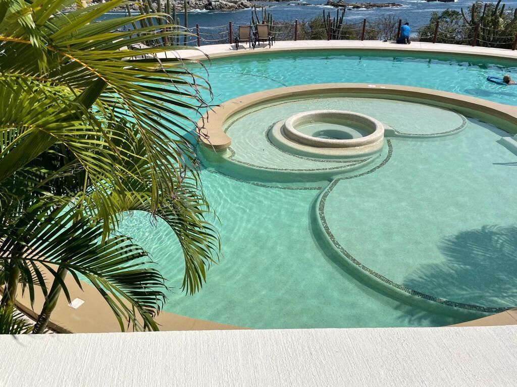 A clear swimming pool with a circular design feature, surrounded by palm trees and a view of the ocean.
