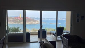 A room with a large sliding glass door leading to a balcony overlooking a coastal area with red buildings and a rocky shoreline. - Oceanview Punta Arrocito Luxury Condo, Near Arrocito Beach (Huatulco)