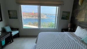 A bedroom with a large bed, a desk, and a chair. There is a window with a view of the sea and a building. - Oceanview Punta Arrocito Luxury Condo, Near Arrocito Beach (Huatulco)