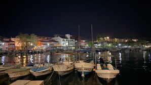 Ein Hafen bei Nacht mit vertäuten Booten, Gebäuden im Hintergrund und einer ruhigen Wasseroberfläche, die Lichter reflektiert.