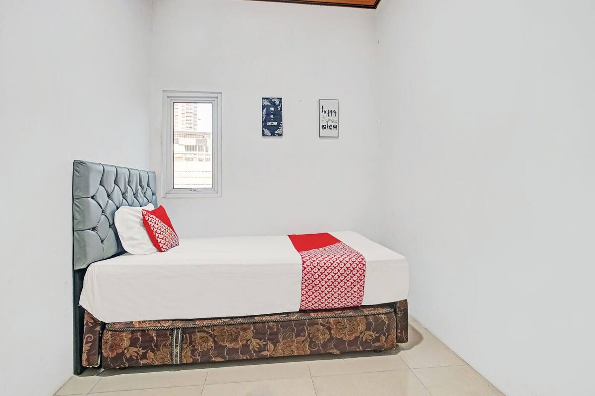 A neatly made bed with a grey headboard, white bedding, and a red throw. There are two wall decorations and a window with a view of buildings.