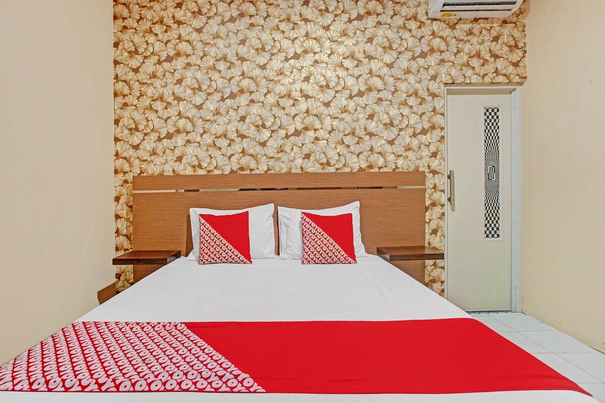 A neatly made bed with a red and white patterned bedspread, a wooden headboard, and a floral wallpaper design on the wall.