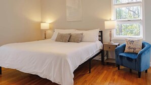 A bedroom with a large bed, bedside lamps, a chair with a cushion, and a window with a view of trees.