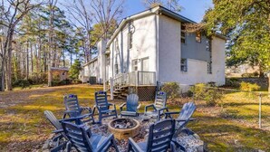 A white building with a porch and a fire pit with chairs in front.