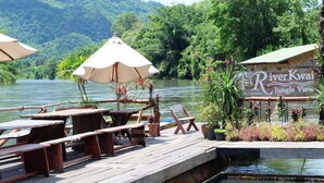 A riverside dining area with wooden tables and chairs, umbrellas, and a sign for 'River Kwai Jungle View'. - River Kwai Jungle View (Sai Yok)