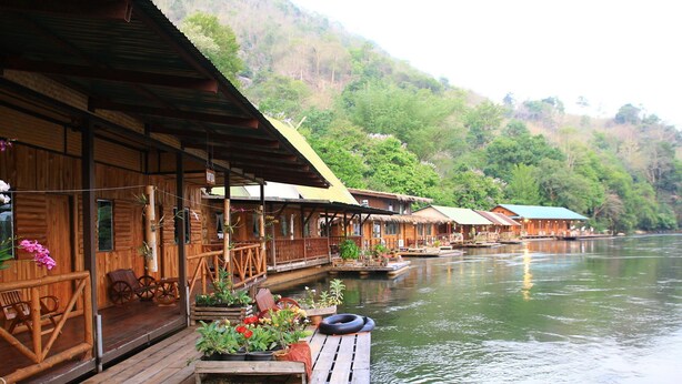 A row of wooden floating houses with thatched roofs, connected by wooden walkways over calm water, surrounded by lush greenery. - River Kwai Jungle View (Sai Yok)