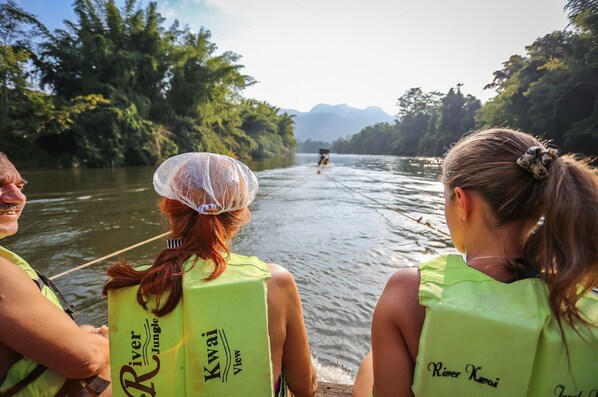 Tres personas en un bote con chalecos salvavidas verdes que tienen texto, rodeadas por un río y vegetación.